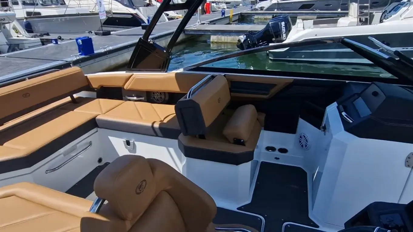 Interior of a modern motorboat featuring tan seating, a steering console, and a view of nearby boats at the marina.